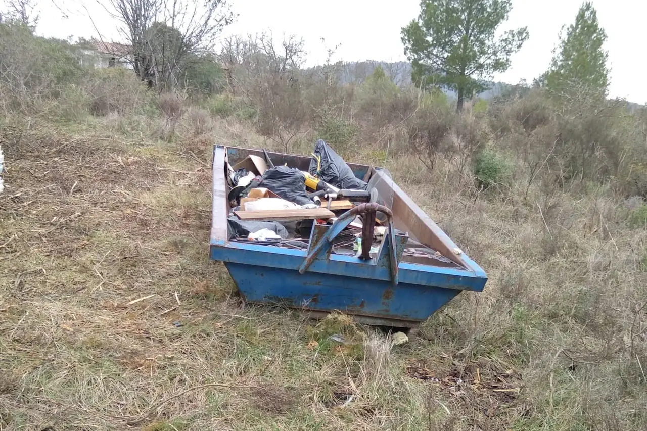 Benne bleue posée sur un terrain en friche, chargée de sacs, de planches et d’encombrants